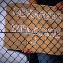Picture of a person behind a chain linked fence holding a sign that says, "Refugees Welcome"